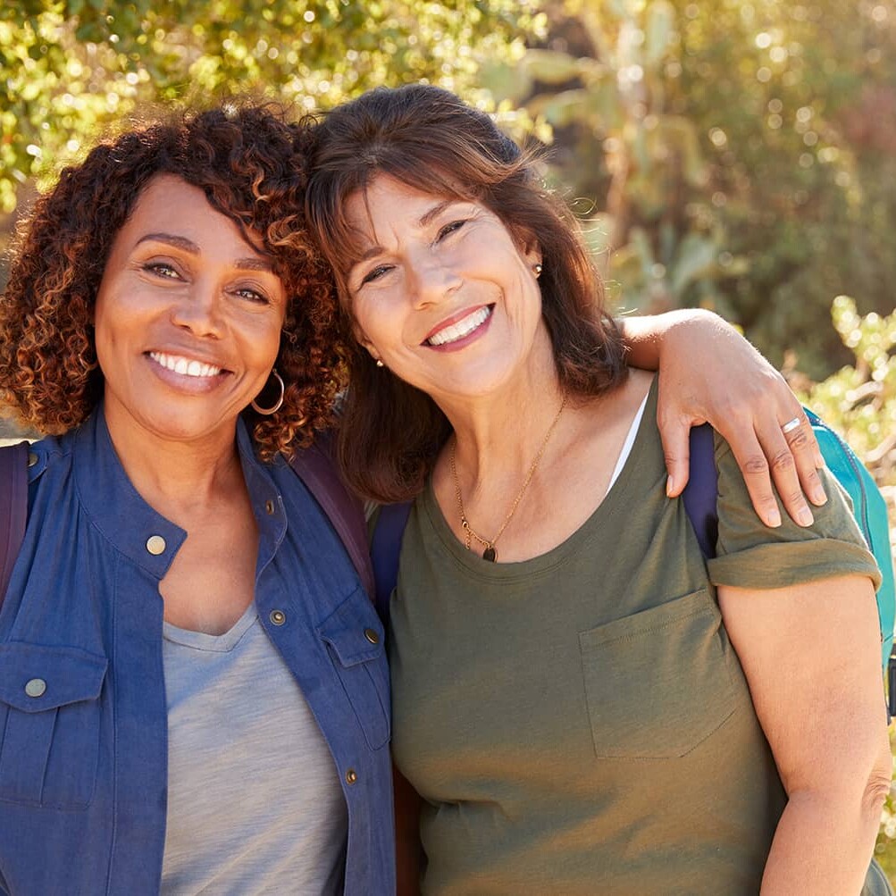 two middle aged woman smiling at the camera in front of trees