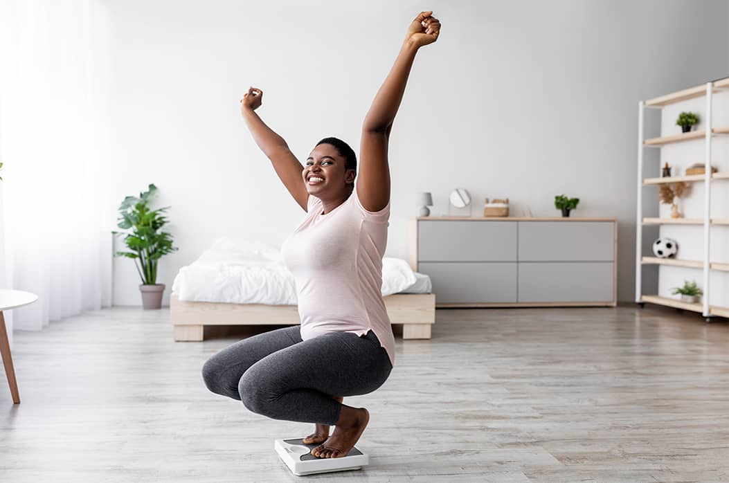 woman in bedroom smiling and raising arms in celebration on scale